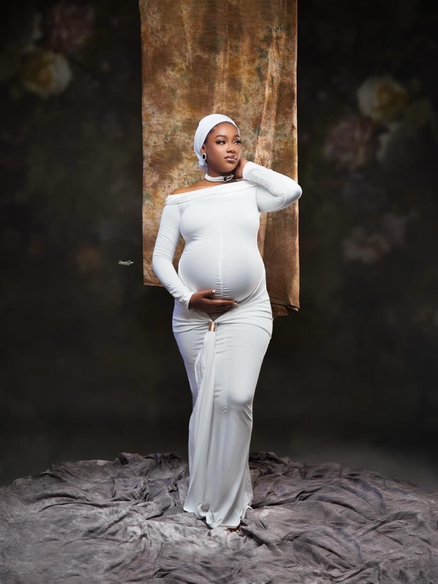 Future maman en robe blanche, mains posées sur le ventre, devant un drapé doré